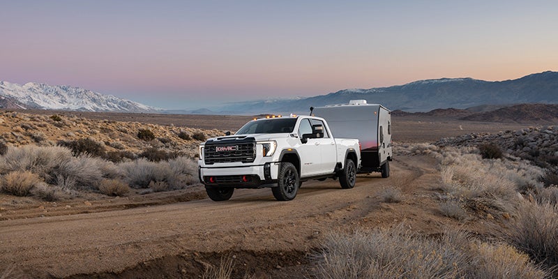 A white 2024 GMC Sierra 3500 hauling a large trailer down a dirt road with the sun setting over mountains in the background