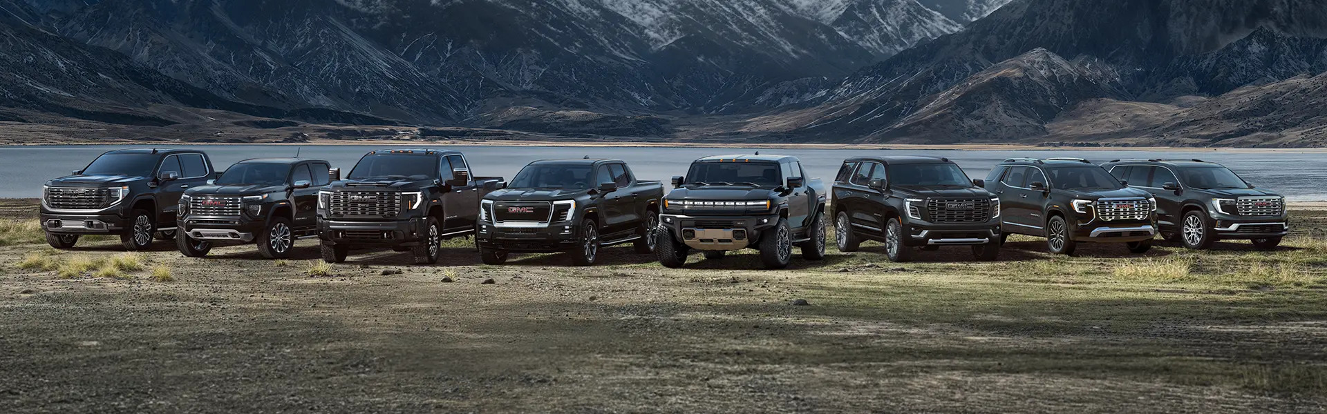 Lineup of GMC trucks and SUVs parked in mountain landscape near a lake.